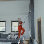 Woman in overalls on a ladder cleaning a high wall in a stylish modern living room.